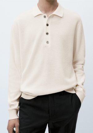 Mand iført en cremefarvet langærmet strikket polo med fire knapper og sorte bukser, stående med den ene hånd i lommen mod en ensfarvet baggrund.