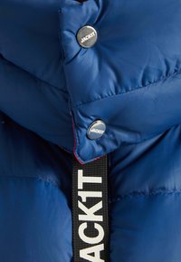 Blauwe pufferjack met een hoge kraag, voorzien van zilveren drukknoopsluitingen en een zwarte band met witte tekst. Glad, glanzend textuur.