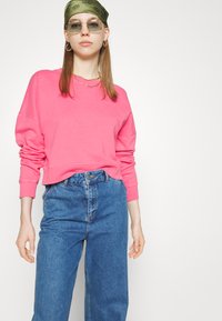Magenta pulover s dolgimi rokavi in rebrastimi manšeti, v kombinaciji z modrimi kavbojkami z visokim pasom. Izrazita barva in sproščen kroj.