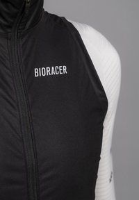 Zwarte fietsvest met hoge kraag, rits aan de voorkant en wit "BIORACER" logo. Heeft textuur witte mouwen met ribbelpatronen.