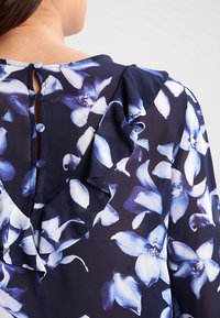 Blomstret bluse med mørkeblå baggrund, med blå og hvide blomsterprint, flæsedetaljer og knaplukning bagpå.