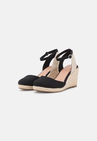 Sandalias de cuña negras de lienzo con un tacón trenzado, parte trasera abierta y correa en el tobillo. La suela presenta una cuerda de estilo espadrille natural.