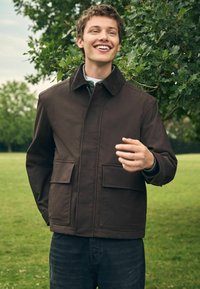 Chaqueta marrón con cuello de pana, cremallera frontal y dos grandes bolsillos de parche. Llevada sobre una camiseta de color claro frente a un fondo verde exuberante.
