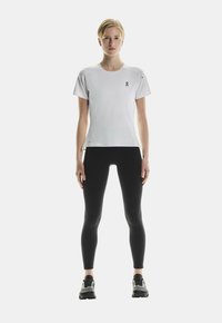 T-shirt d'athlétisme blanc à manches courtes avec un logo noir sur la poitrine, associé à des leggings noirs et des chaussures de course noires. Design simple.