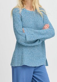 Femme portant un pull en tricot bleu clair et un pantalon bleu, debout les bras croisés devant un fond uni.