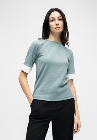 Γυναίκα με σκούρα μαλλιά που φοράει γαλάζιο ριμπ μπλουζάκι με κοντά μανίκια και λευκές μανσέτες, καθώς και μαύρο παντελόνι, στέκεται με το ένα χέρι στην τσέπη.