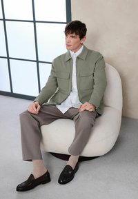 Homme assis sur une chaise blanche arrondie portant une veste vert clair, une chemise à rayures, un pantalon taupe, des chaussettes beiges et des mocassins noirs près d'une fenêtre.
