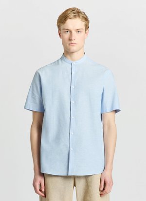 Jeune homme portant une chemise bleu clair à manches courtes avec col officier et un pantalon beige, debout devant un fond uni.