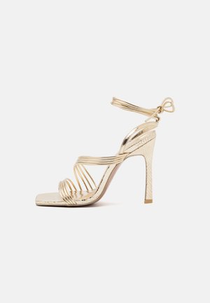 Złote sandały na wysokim obcasie z wieloma spiralnymi paskami, kwadratowym noskiem i teksturowanym obcasem. Oferują cienki pasek wokół kostki z dekoracyjnym skrętem.