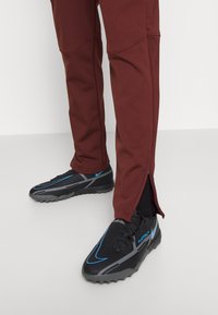 Zwarte sportieve schoenen met blauwe accenten en een getextureerd oppervlak, gecombineerd met bordeauxrode tapered pants met ritsen bij de enkels.