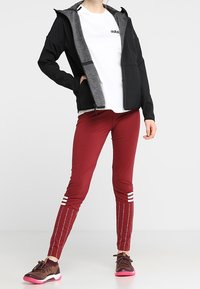 Svart zip-up jacka med grå detaljer, vit t-shirt med logotyp, vinröda leggings med sidostråkar och bruna sneakers med rosa sulor.