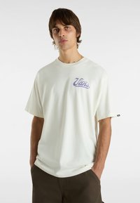 Camiseta gráfica de color crema claro hecha de algodón, con un logo de Vans en púrpura en la parte izquierda del pecho. Manga corta y corte relajado.