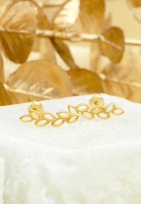 Orecchini d'oro a forma di foglie d'olivo collegate, appoggiati su una pietra bianca texturizzata, con uno sfondo sfocato di foglie dorate.