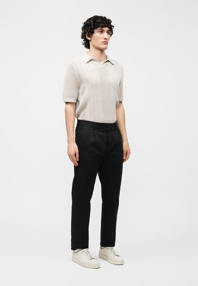 Homme debout portant un polo à manches courtes beige texturé, un pantalon noir et des baskets blanches, faisant face à l'avant sur un fond clair uni.