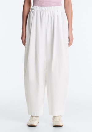 Žena nosí voľné biele nohavice s elastickým pásom, v kombinácii s bielymi teniskami a ružovým pruhovaným topom.