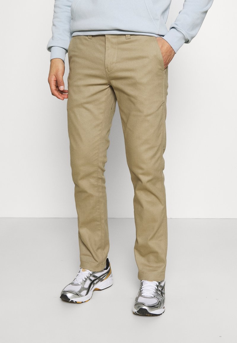 Beige bavlněné chinos s rovný střihem. Má standardní pás a boční kapsy, doplněné bílými a stříbrnými sportovními teniskami.