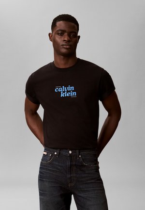 Joven con una camiseta negra de Calvin Klein con el logo en azul y vaqueros oscuros, de pie con las manos detrás de la espalda.