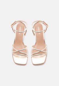 Ζευγάρι μεταλλικές χρυσές σανδάλες ανοιχτές στο δάχτυλο με λεπτούς διασταυρούμενους ιμάντες και αγκράφες στον αστράγαλο, με επίπεδες τετράγωνες σόλες, σε λευκό φόντο.