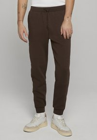 Bruine joggingsbroek met een elastische tailleband en trekkoord, met witte zijstrepen, aansluitende boorden en een gladde textuur. Gecombineerd met witte sneakers.