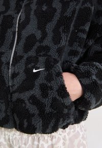 Fleece jakna v črno-sivi leopardski vzorček z zadrgo in Nike logo; ima teksturirano blago in rebraste manšete.
