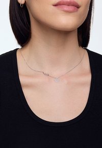 Srebrny naszyjnik z małym skrzydłem i zawieszką w kwiatowy wzór na delikatnym łańcuszku, noszony z czarnym t-shirtem.