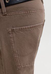 Pantalon en denim marron en gros plan montrant une poche arrière et une ceinture noire passée dans la ceinture.