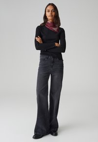 Schwarzes geripptes Langarmoberteil, hoch taillierte dunkelgraue weit geschnittene Jeans und ein bordeauxroter Schal mit weißen Streifen. Glatte Texturen und taillierter Schnitt.