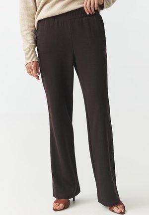 Person, die schwarze Hosen mit weitem Bein, einen beigen Pullover und braune offene High Heels trägt, steht vor einem schlichten Hintergrund.