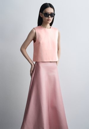 Žena oblečená v bezrukávovom bledoružovom topu a zodpovedajúcej dlhej sukni, s čiernymi slnečnými okuliarmi a rovnými tmavými vlasmi, stojaca pred sivým pozadím.