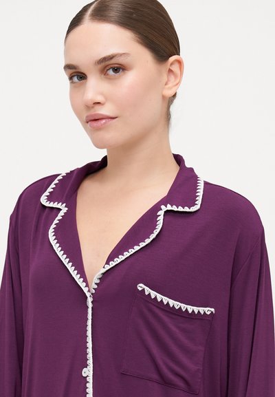 Cămașă de pijama mov dintr-un material moale, având un guler alb cu bordură, o deschidere cu butoane în față și un buzunar cu detalii decorative.