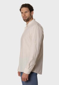 Camisa de lino color beige claro con cuello alto, mangas largas y detalles de botones, presentada ajustada de manera ordenada en un modelo masculino sobre un fondo neutro.