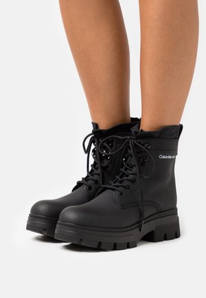 Svarta ankelstövlar av gummi med snörning, vadderad krage och tjock, räfflad sula för extra grepp och stabilitet.