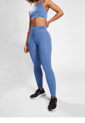 Blå træningssæt bestående af en tætsiddende crop top og højtaljede leggings. Glat tekstur med logoer, parret med sorte sportsko.