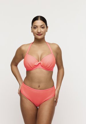 Vrouw die zelfverzekerd staat in een koraalkleurige, gestructureerde halterbikini met knoopdetail op het topje, tegen een effen witte achtergrond.