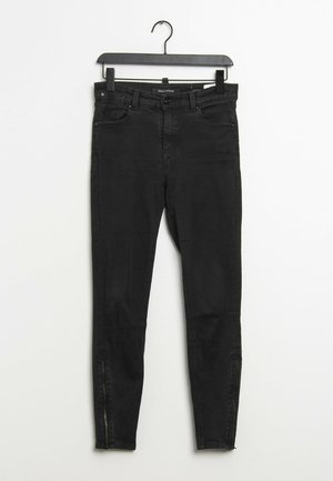 Black slim-fit jeans with front pockets and ankle zippers hanging on a black plastic hanger against a white wall.