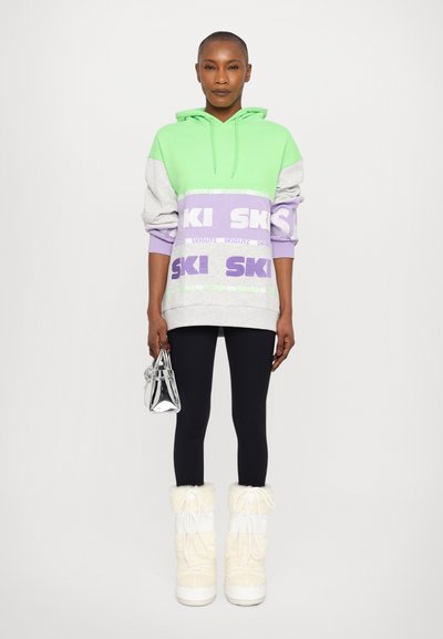 Hooded sweatshirt in green, gray, and lavender with "SKI" printed. Black leggings, white furry boots, and a small silver bag. Smooth texture.