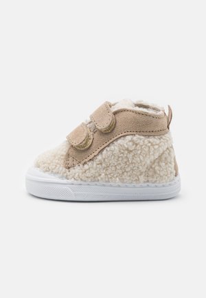 Chaussure beige pour tout-petit avec tissu blanc duveteux, deux bandes Velcro et semelle en caoutchouc blanc sur fond blanc uni.