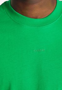 Gros plan sur une personne portant un t-shirt vert vif à col rond avec "SANDRO" brodé en petites lettres blanches sur la poitrine.