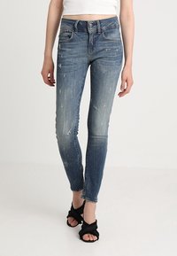 Vaqueros ajustados en denim azul desvaído con detalles desgastados y una cintura de media altura. Presentan un estilo clásico de cinco bolsillos y cierre de cremallera.