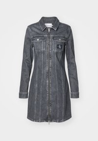 Denimkjole i mørkegrå farge, lange ermer, frontglidelås og to brystlommer. Har en CK-logo-patch og vertikale sømmer.