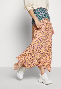 Falda maxi con base amarilla, decorada con patrones florales en púrpura y rosa. Cintura ajustada en verde de contraste. Combinada con zapatillas blancas.