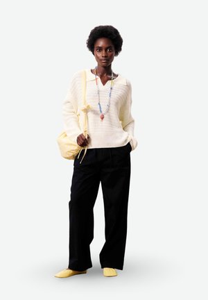 Femme portant un pull en maille crème, un pantalon noir à jambes larges, des chaussures jaunes, un collier coloré en perles et portant un sac à bandoulière jaune.