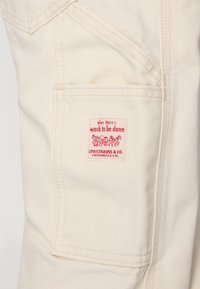 Tejido beige con una superficie texturizada, que presenta una etiqueta roja y blanca que dice "cuando hay trabajo por hacer" y "LEVI STRAUSS & CO."