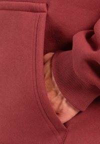 Bordowy sweatshirt z kieszenią kangurzą, ściągaczami u dołu rękawów oraz miękkim, fakturowanym materiałem. Widać szwy wokół kieszeni.