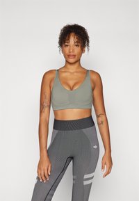 Sutiã desportivo em verde oliva, com um decote em V, alças largas e perfurações subtis. Combinado com leggings cinzentas texturizadas com riscas.