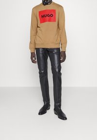 Persona vistiendo una sudadera beige con el logo rojo de HUGO, pantalones de cuero negro, zapatos de cuero negro y un pañuelo estampado en el cuello.