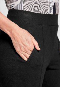 Zwarte broek met een gladde textuur en een elastische tailleband, voorzien van zijzakken en een getailleerde pasvorm. Detail van een hand die in een zak rust.