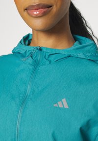 Giacca leggera teal con zip e cappuccio, caratterizzata da una finitura texture e un logo grigio visibile sul petto.
