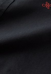 Tessuto nero con una trama testurizzata, caratterizzato da un piccolo logo ricamato rosso nell'angolo. Morbido al tatto, mostrato in posizione piegata.