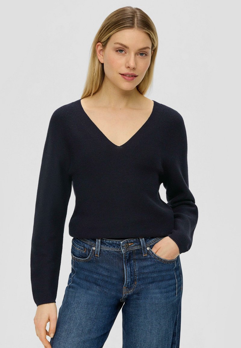 Marineblauer V-Ausschnitt-Pullover mit langen Ärmeln, aus einem strukturierten Strickstoff, kombiniert mit eng anliegenden blauen Jeans.